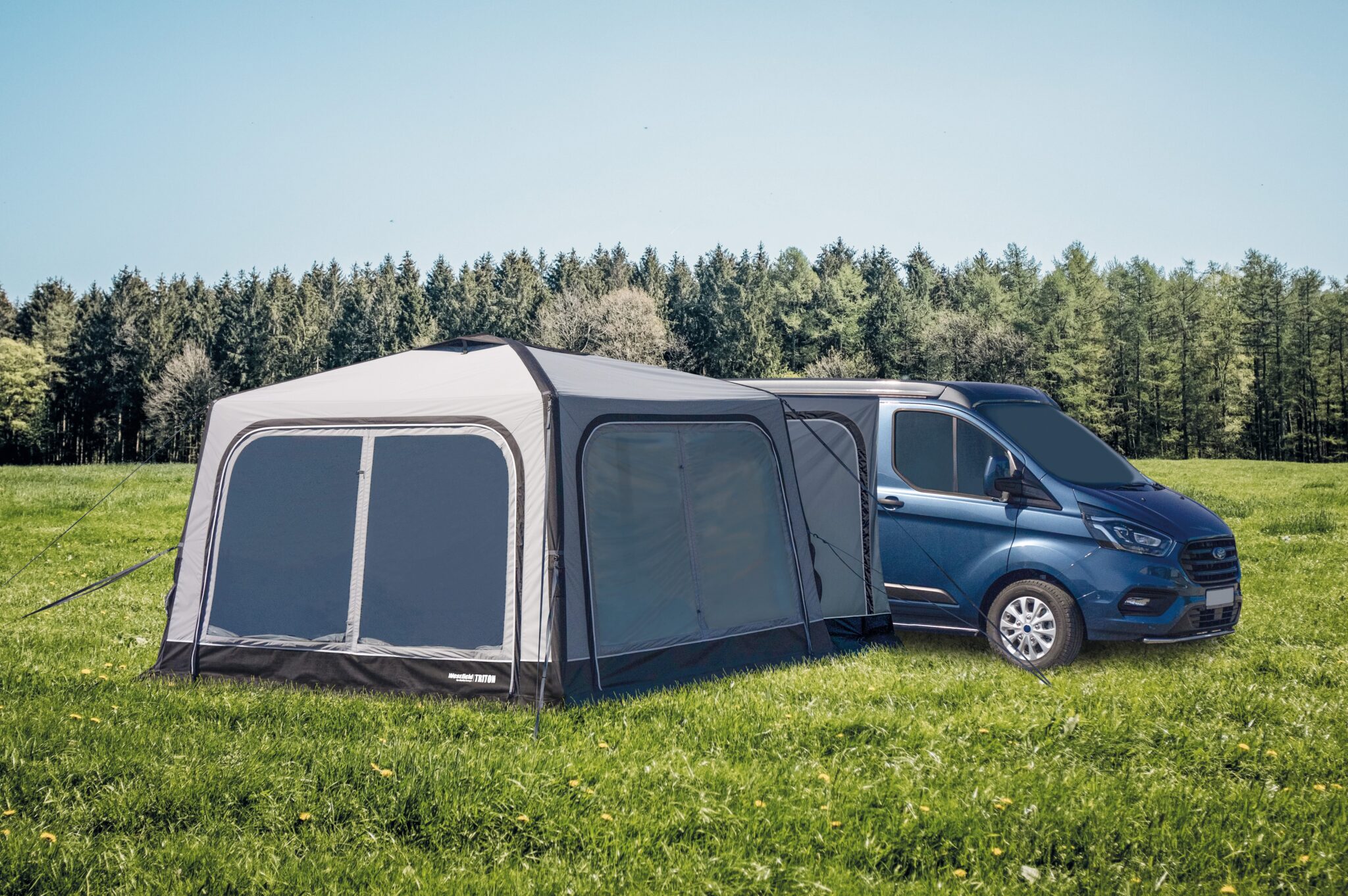 Campervan mit angeschlossenem Zelt auf Wiese vor Wald.